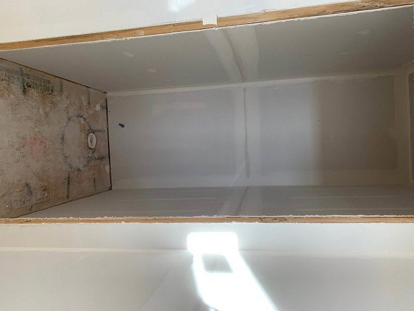 White plaster bathroom wall with a rectangular hole, sunlight streaming through window, wood framing visible, person painting wall nearby