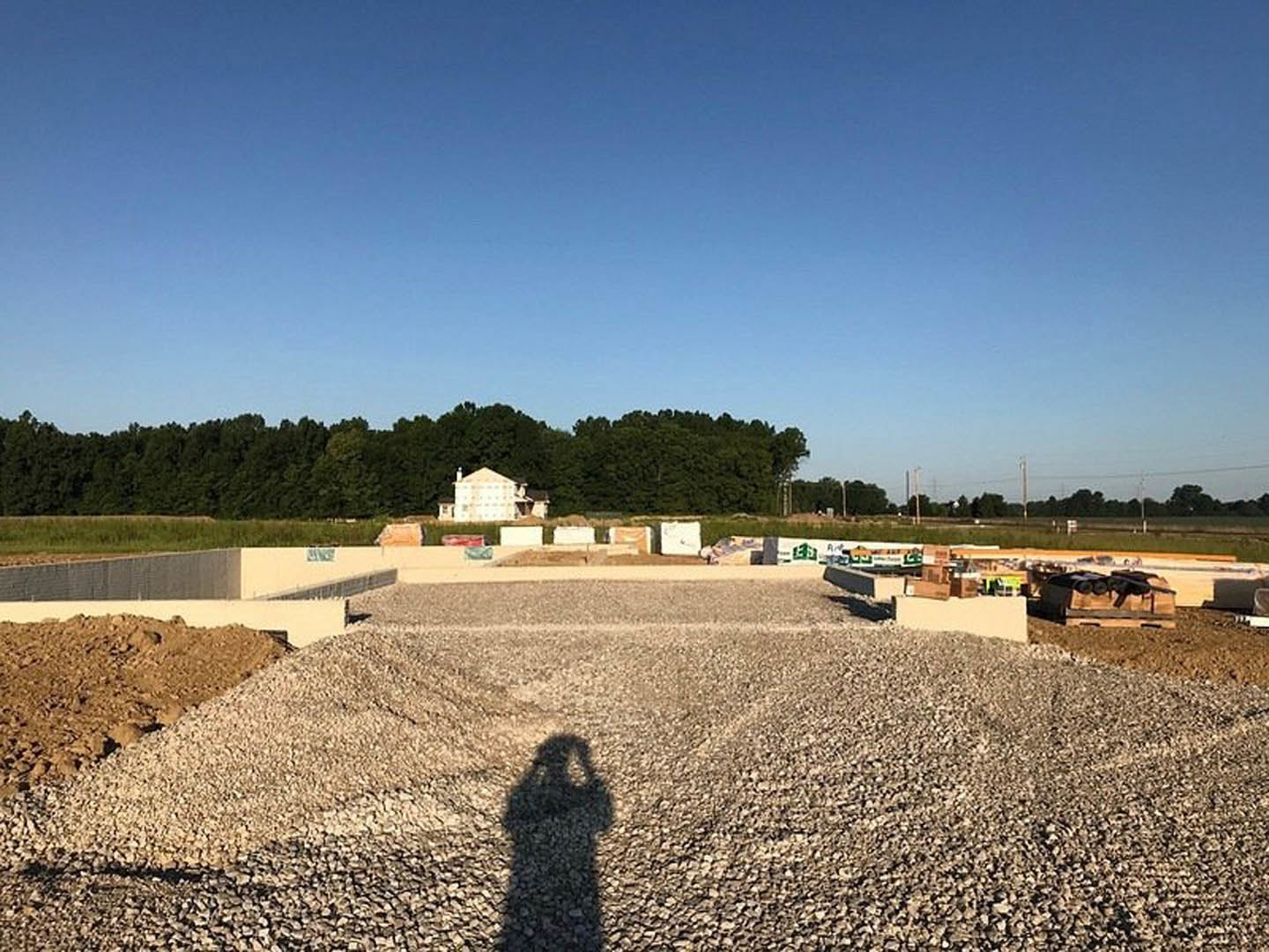 Shadow of a person cast on a gravel driveway with a blurry house partially visible among trees under a clear blue sky