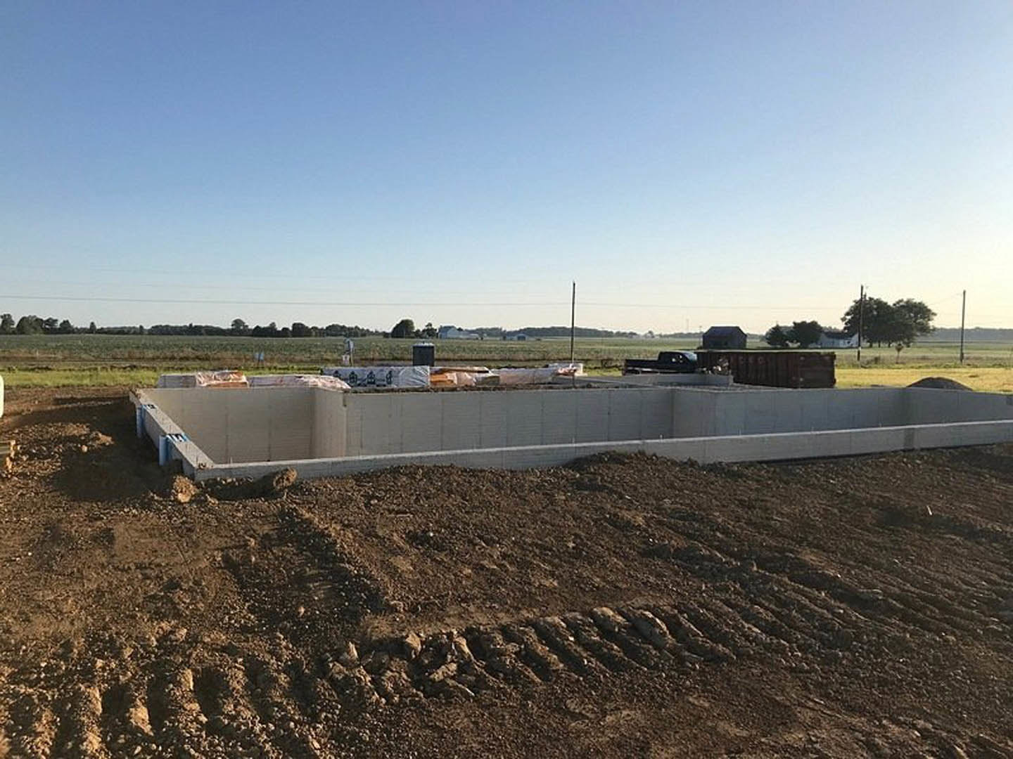 Concrete foundation and retaining wall surrounded by dirt and soil, open excavation area, grassy field, trees in background, blue sky with scattered clouds