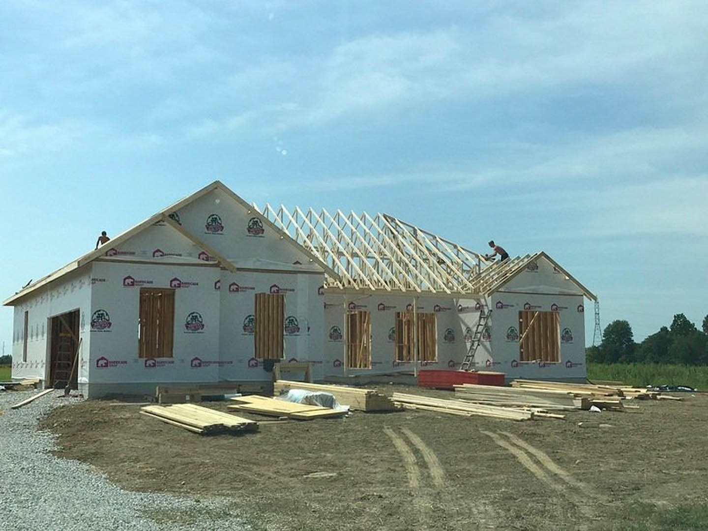Wood-framed house under construction with two people working on the roof, exposed beams, unfinished exterior, and ladder leaning against the structure