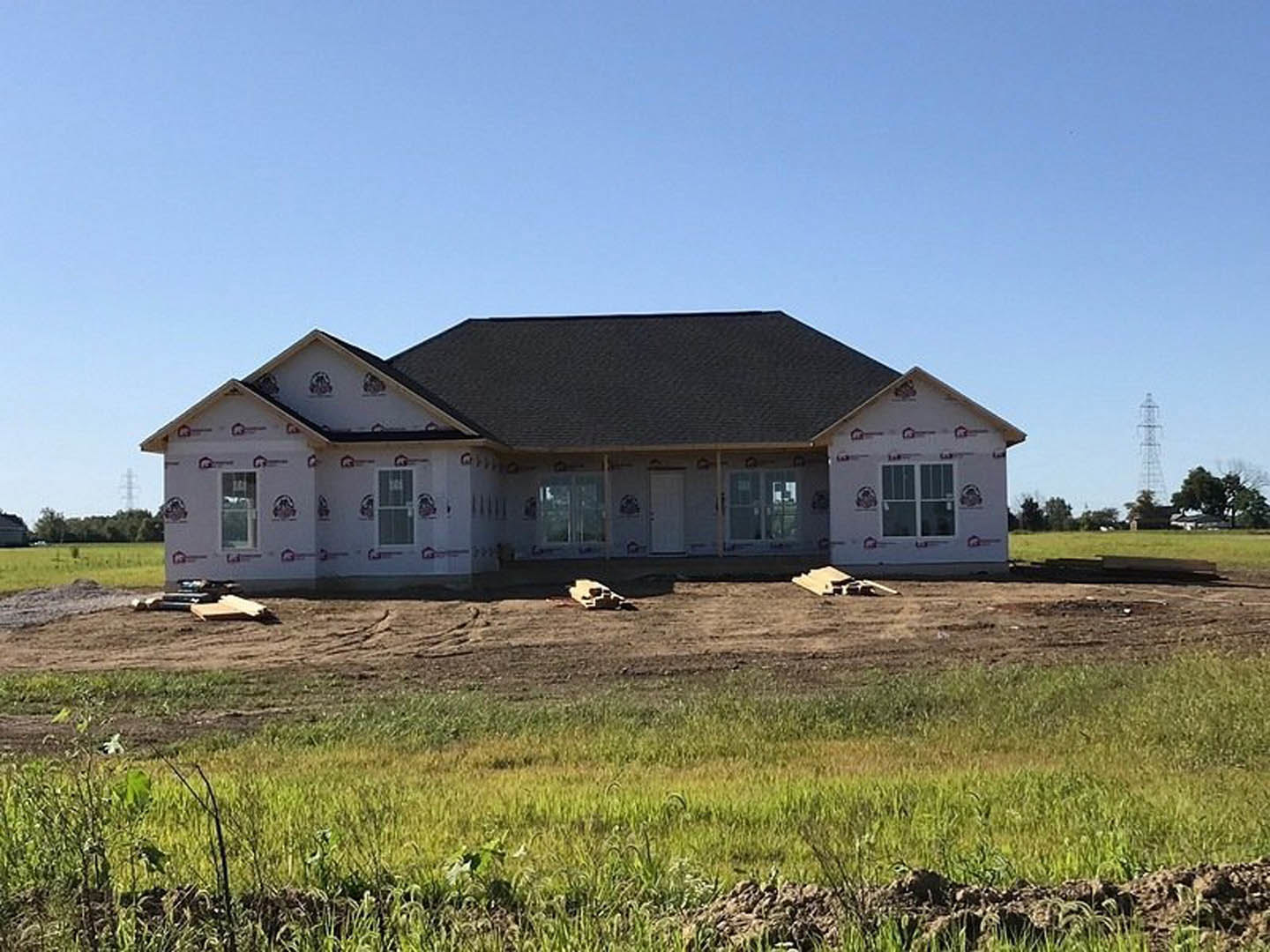 Partially built house with exposed wood framing, black shingle roof, surrounded by green grass and open land under a clear sky