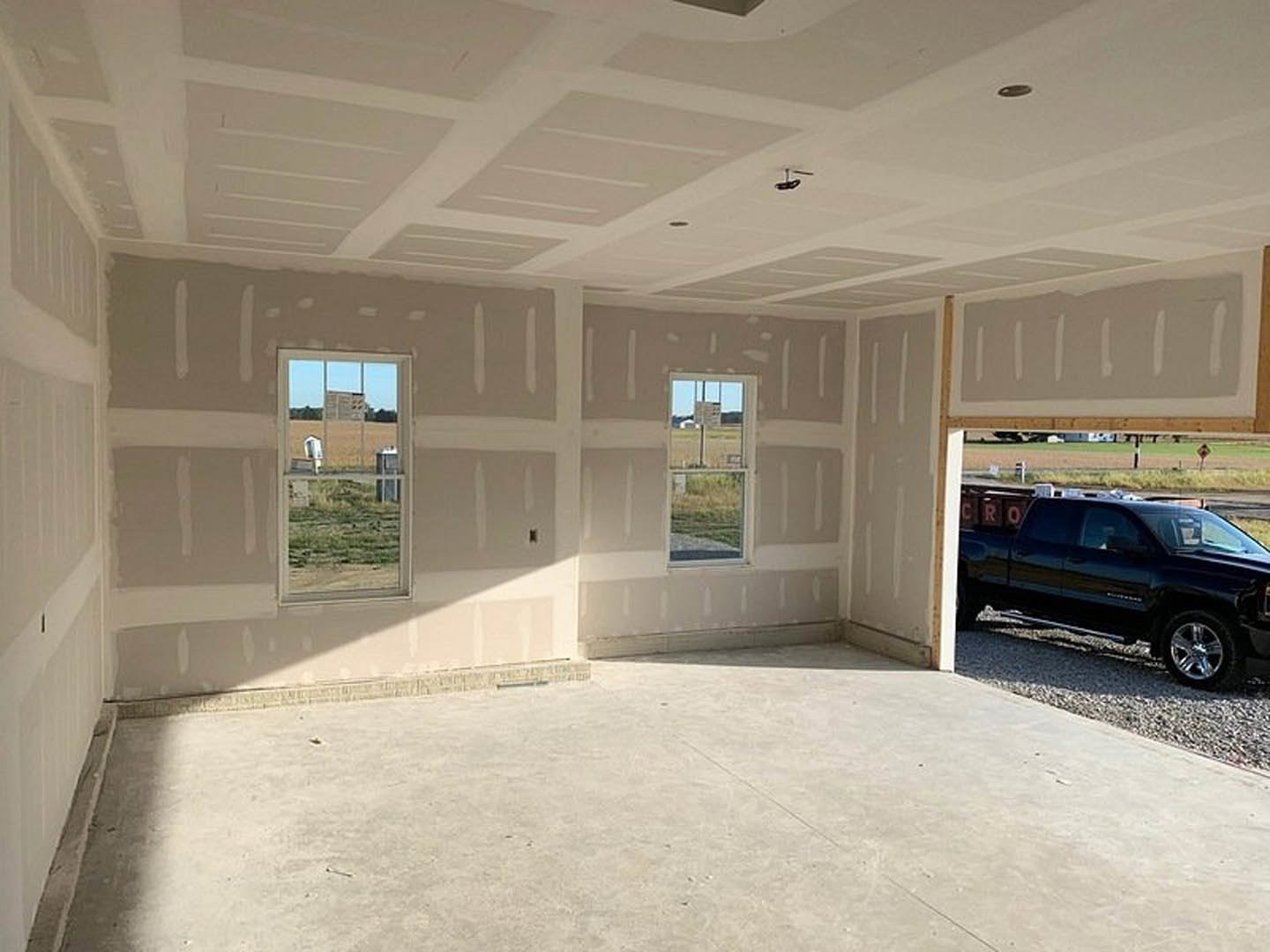 White-walled room with large windows, concrete floor, sunlight casting shadows, black truck parked on gravel outside, field and fence visible through window, close-up of car tire