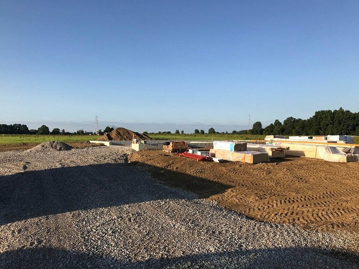 Gravel-covered construction site with stacked boxes, plastic-wrapped bags, scattered equipment, clear blue sky, and distant trees