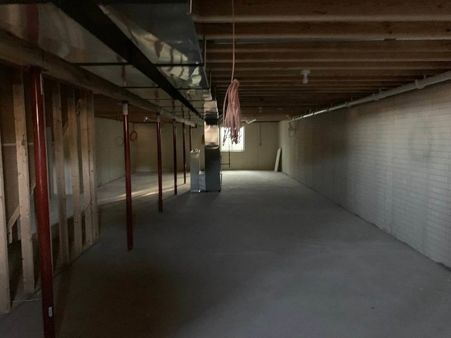 White brick wall and ceiling in a basement room with concrete floor, red support poles, exposed light bulb, and a blurry pink object in the foreground.