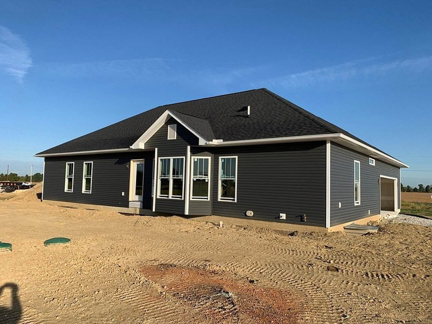 Partially built house with black roof and white-framed window, surrounded by sandy dirt field under cloudy sky