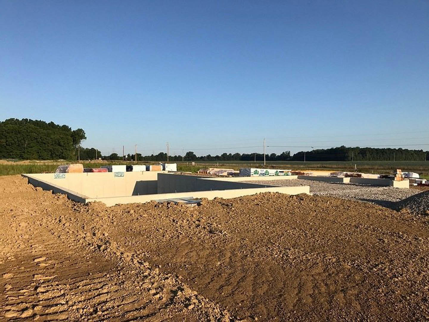 Partially built custom home surrounded by piles of dirt, concrete blocks, and mature trees under a blue sky