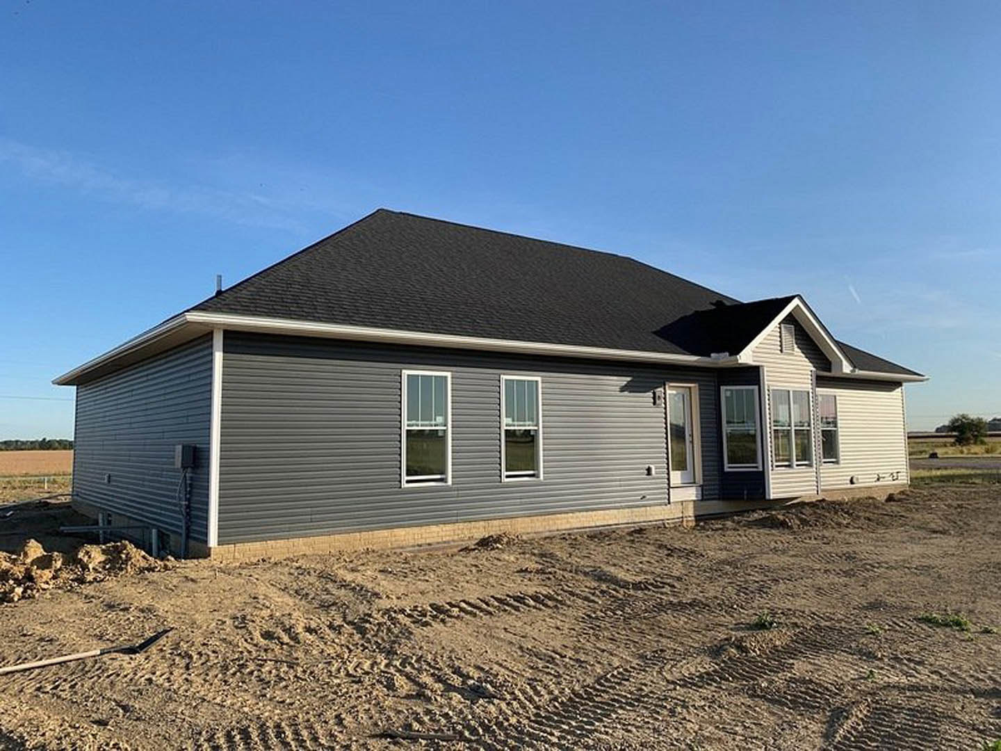 Partially built house with gray shingle roof, white-framed windows, sandy ground with tire tracks, exposed siding, and clear sky overhead