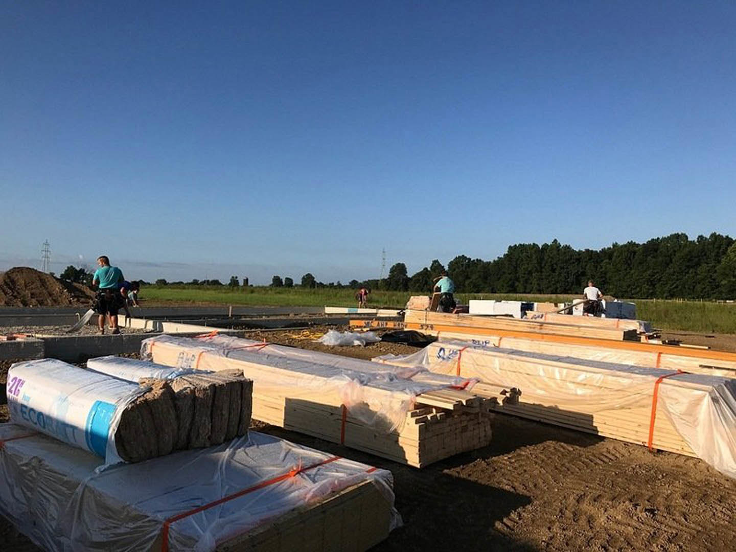 Modern farmhouse exterior with natural wood beams, white siding, and large windows, set against a grassy field under a blue sky with scattered clouds.
