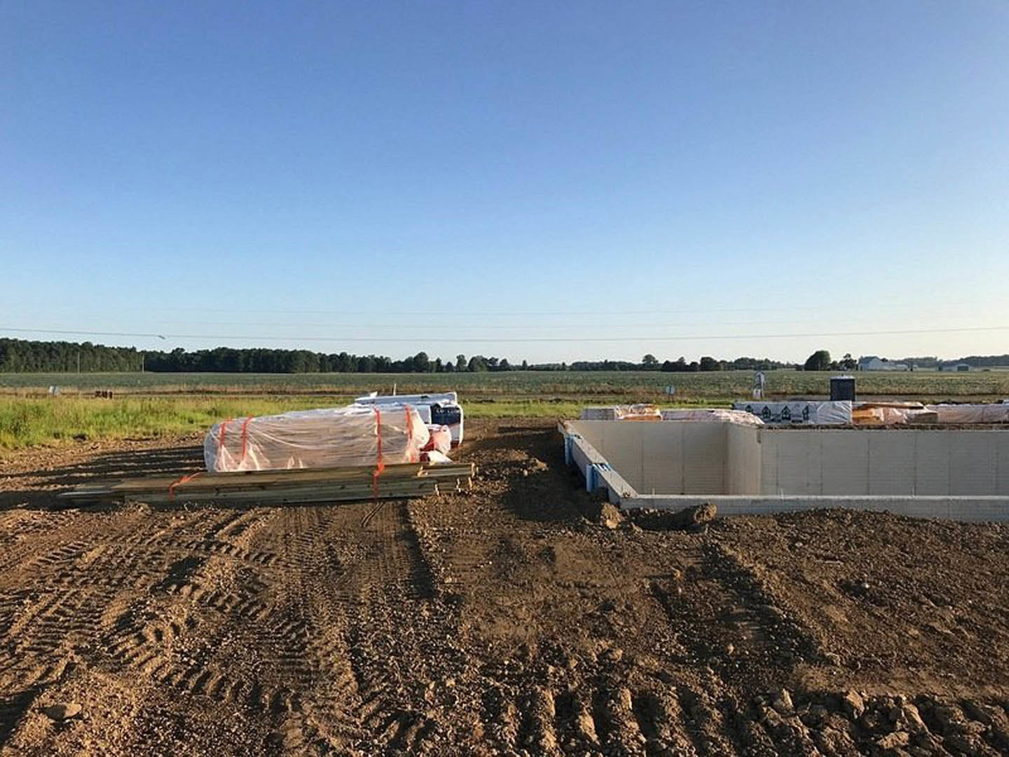 White exterior wall under blue sky, dirt ground with piles of sand and wood, plastic-wrapped lumber, scattered construction materials, man standing near grassy field.