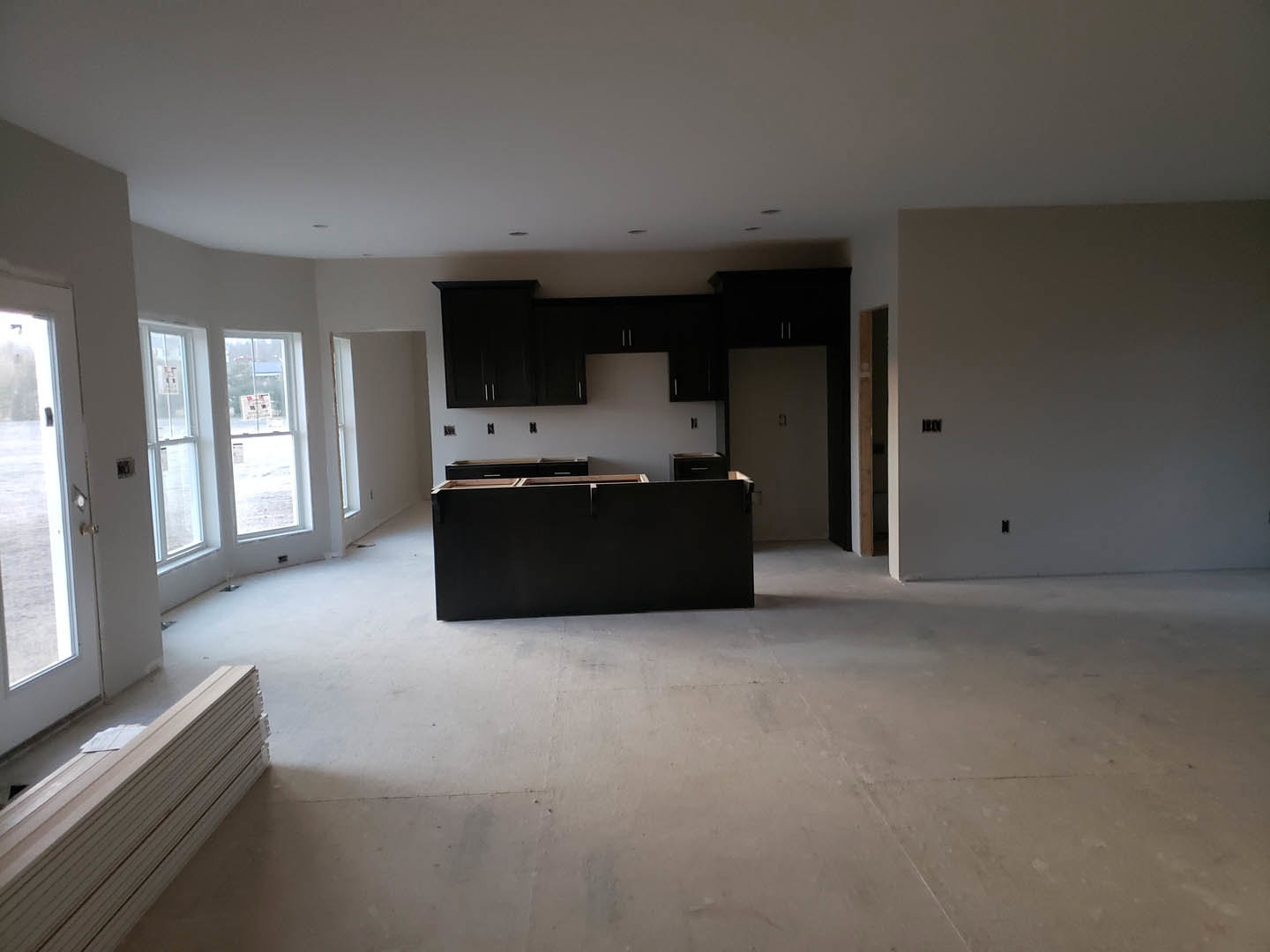 Open-concept kitchen with matte black island topped in light wood, concrete flooring, plaster walls with light switches, large window, and stacked wood planks near a doorway