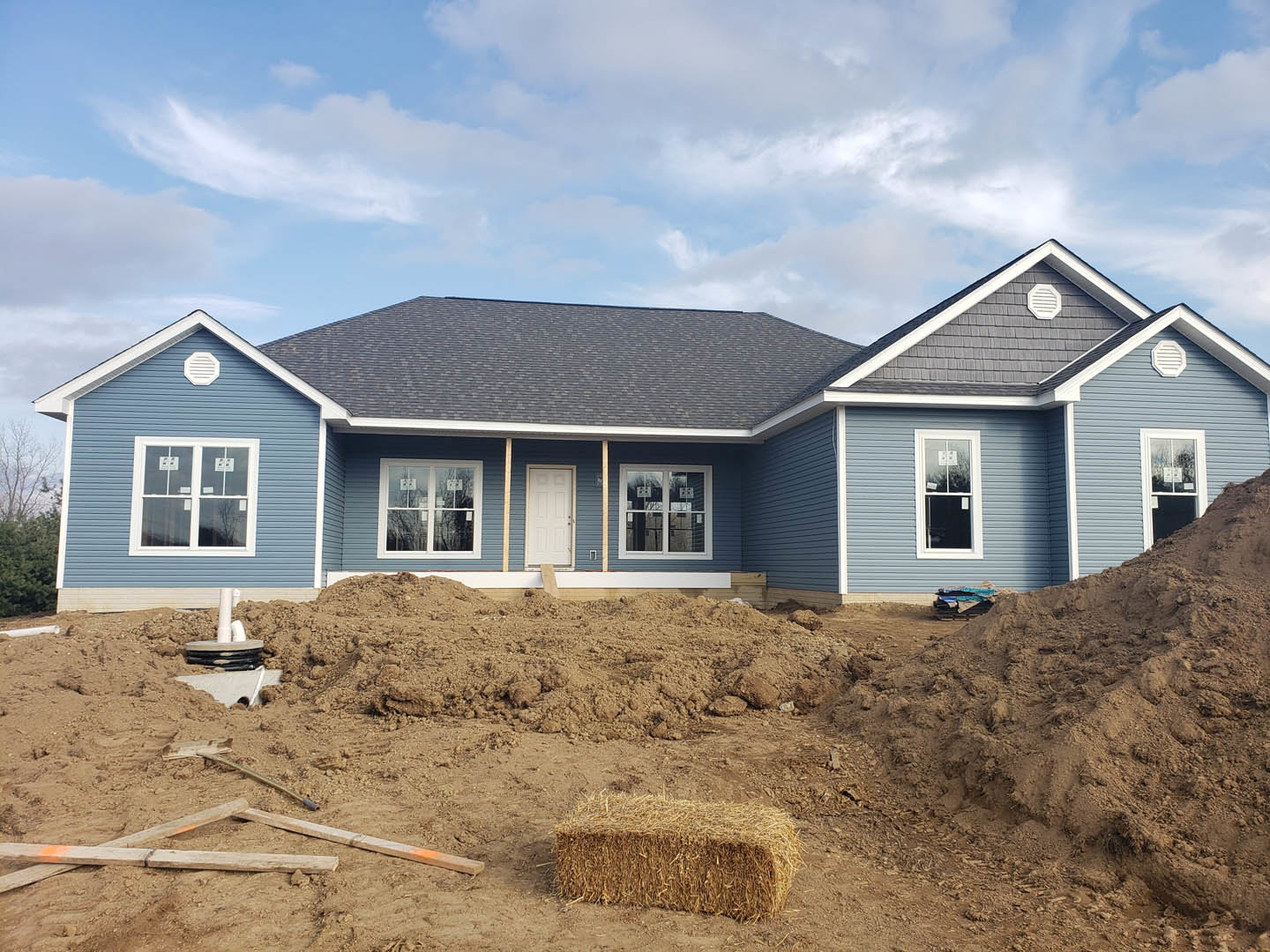 Two-story house with white door and windows, dirt yard with large pile of soil in front, cloudy sky overhead