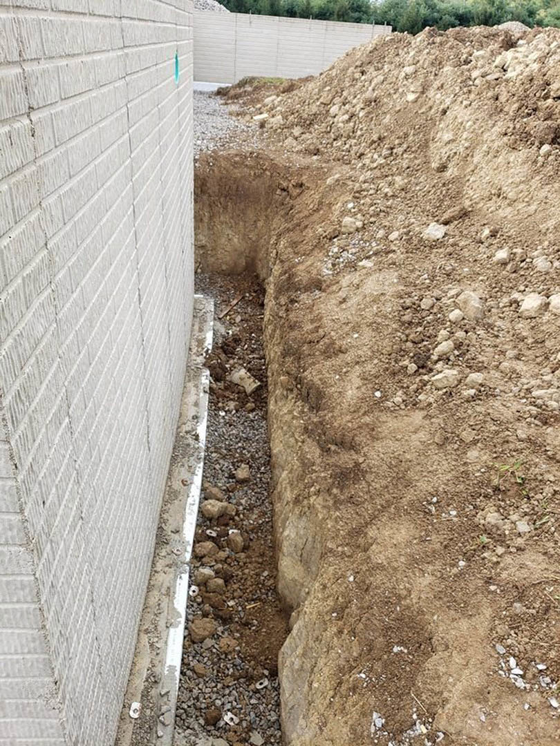 Freshly dug trench in compacted dirt, bordered by rocks and gravel, with a white brick wall and trees in the background, pile of soil near a wooden fence