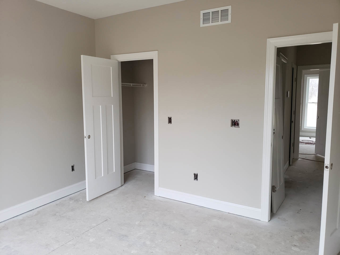 White-walled room with white flooring, ceiling vent, and a white door featuring a round hole, adjacent to a hallway with a window.