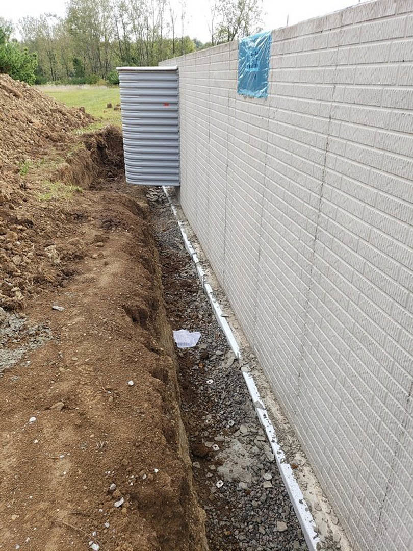 White brick exterior wall with a large hole in the dirt and gravel ground, blue plastic sheeting partially covering bricks, nearby soil and sparse plants visible.