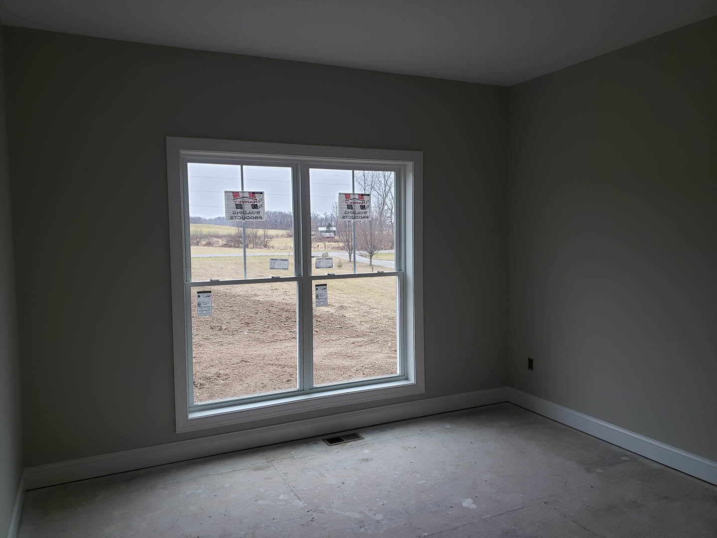 Bright room with plaster walls, large window overlooking dirt field, floor vent centered, window adorned with multiple signs containing text.