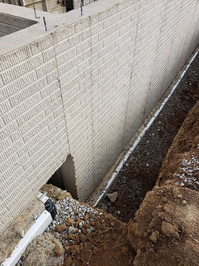 White composite exterior wall with a rough-edged hole, black and white work glove visible near opening, ground surface and plastic strip at base.