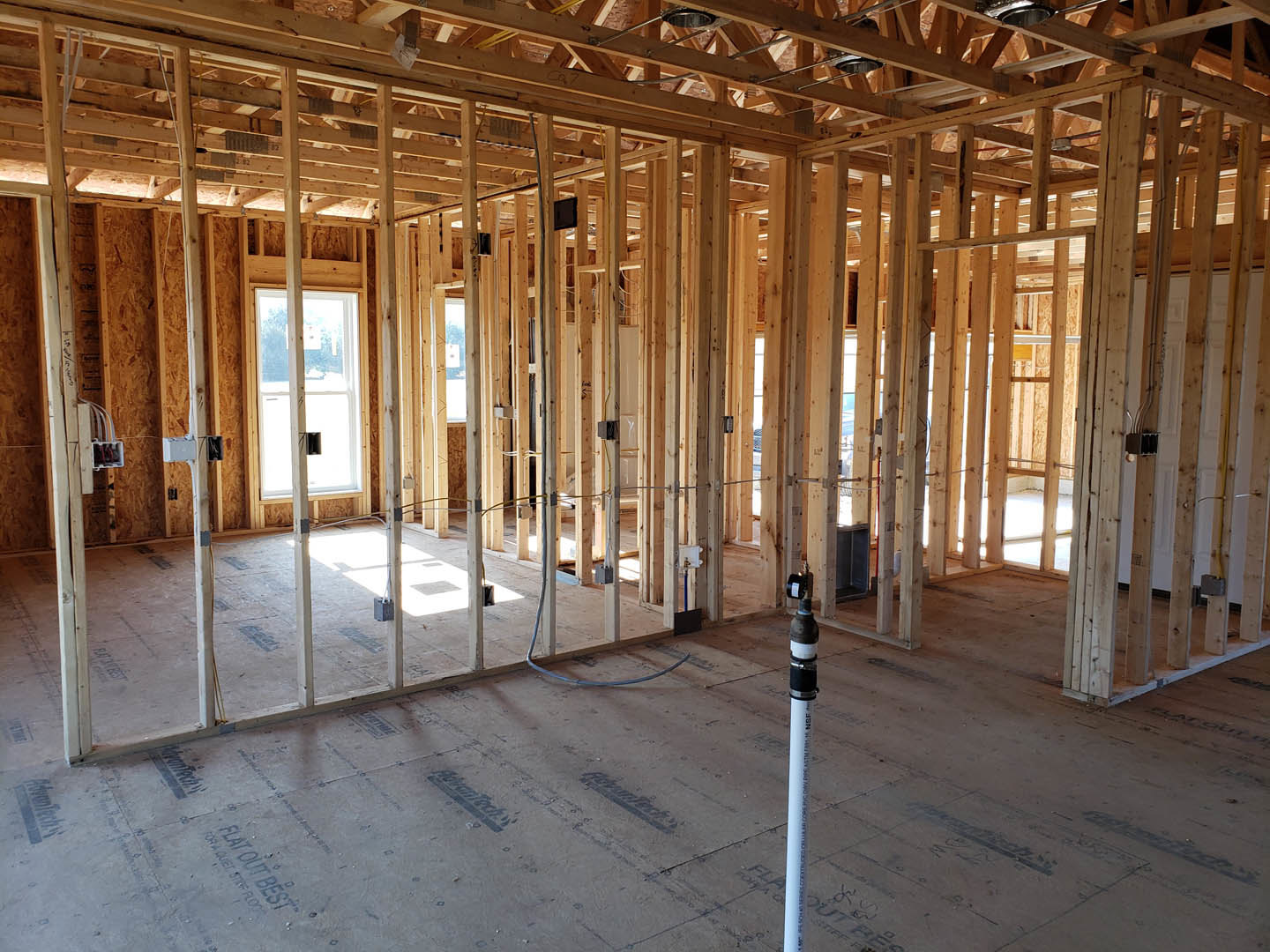 Wood-framed room under construction with exposed beams, white-framed window, unfinished ceiling, and visible white pipe with black text