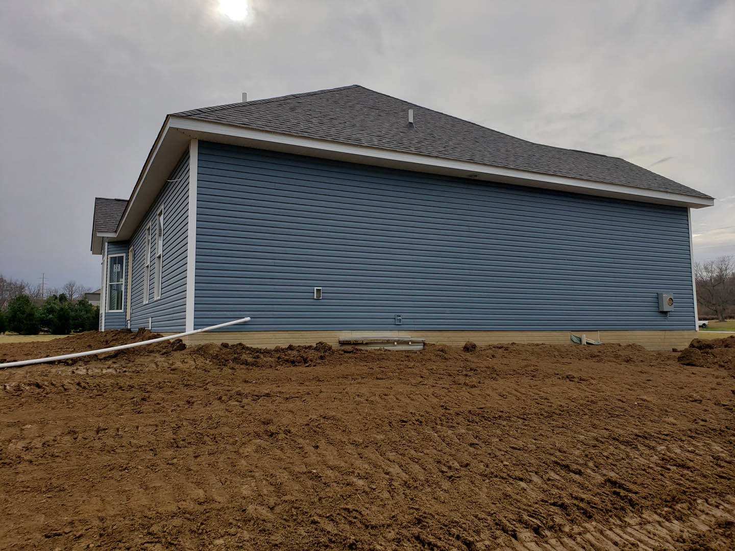 Blue siding on a house with exposed dirt foundation, white pipe emerging from soil, open field in foreground, distant trees and cloudy sky in background