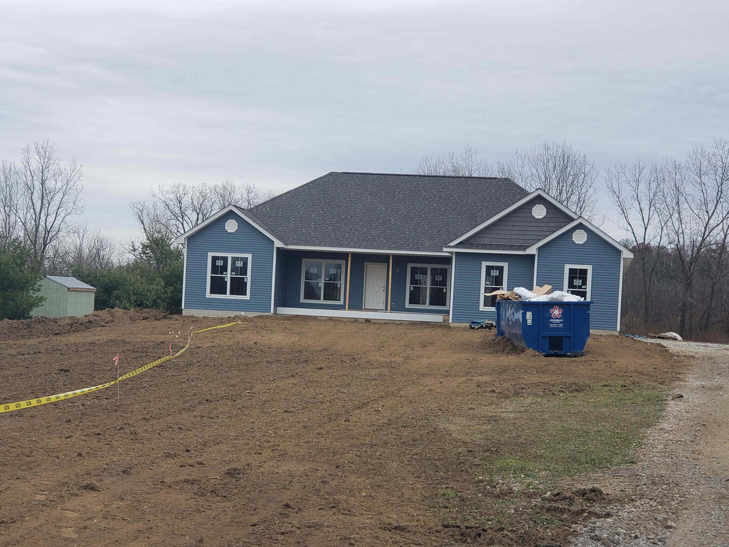 Two-story house with blue metal roof, blue siding, white door, blue dumpster filled with debris on dirt yard, yellow caution tape on hillside, cloudy sky and trees in background