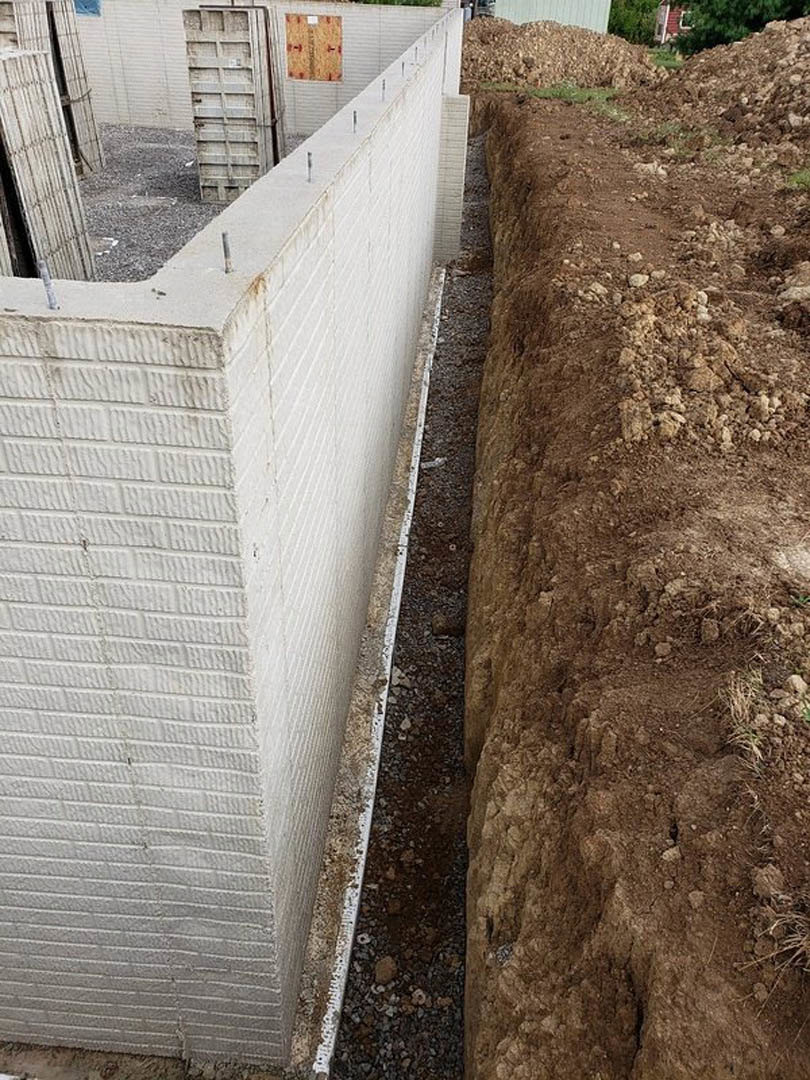 White brick exterior wall with cement base, brown soil trench running along the ground, sparse plants visible near concrete foundation