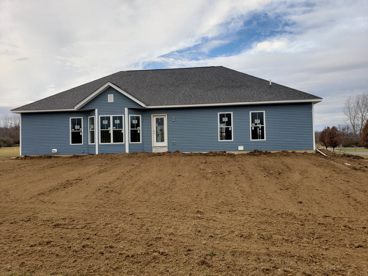 Blue house with white trim and white door, glass-paneled entry, dirt field in foreground, cloudy sky overhead, sparse trees in background.