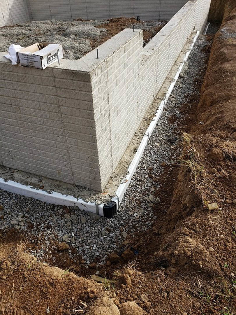 Red brick exterior wall with a hole in the ground, exposed concrete foundation, pile of soil, and a cardboard box nearby