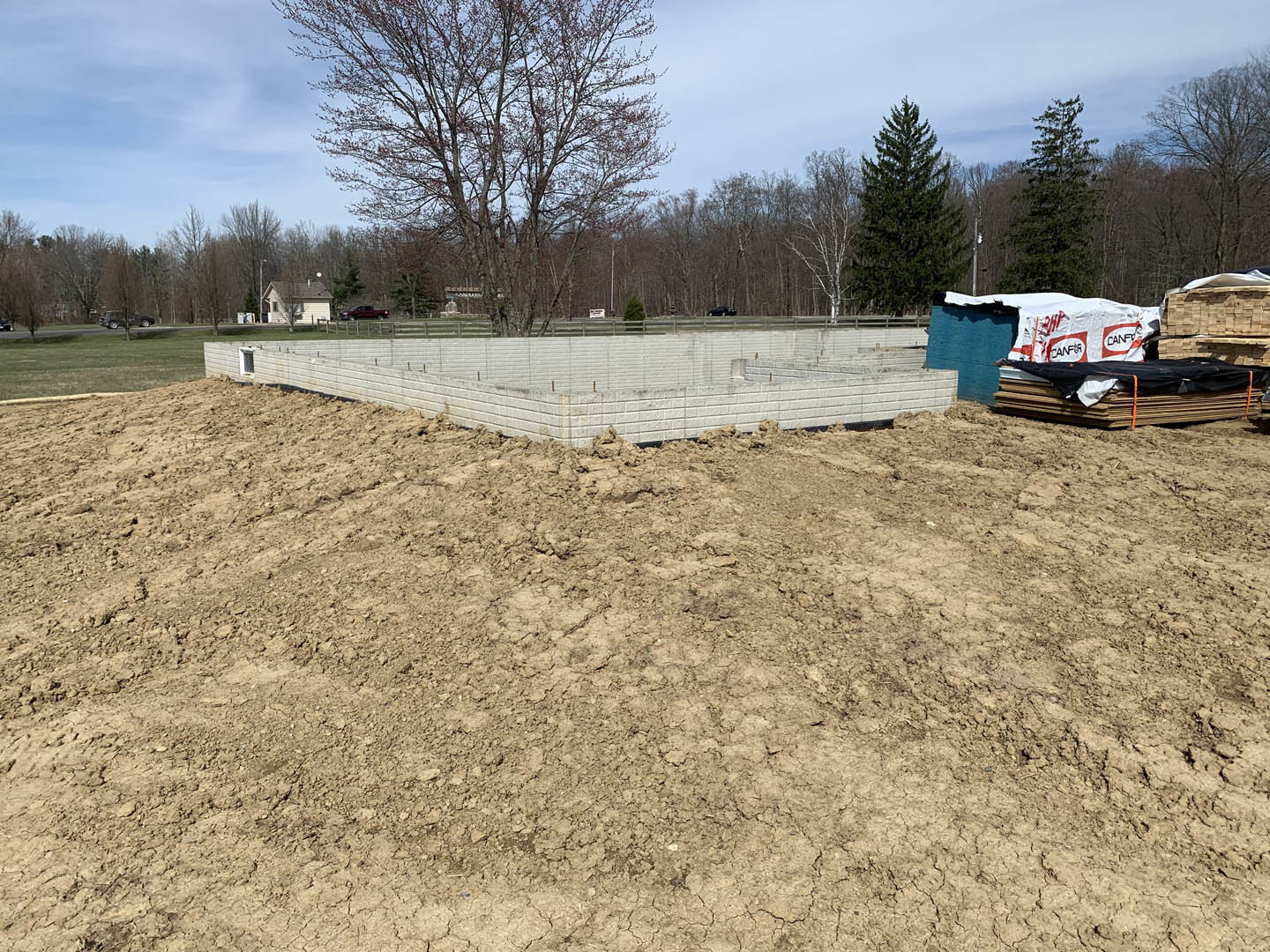 Dirt field bordered by a wooden fence, mature trees in the background, white tarp covering stacked wood near a concrete wall