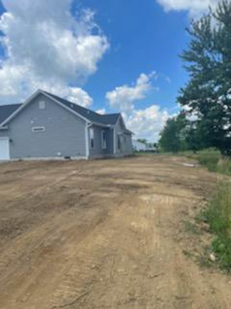 Single-story cottage with light siding, surrounded by mature trees, set along a dirt road under a blue sky with scattered clouds