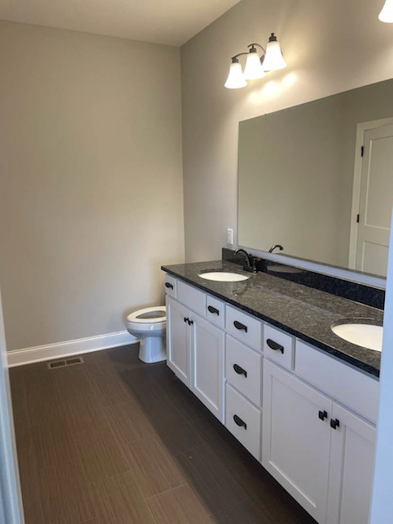 Bathroom with white cabinetry featuring black handles, undermount sink with chrome faucet, white toilet, wood flooring, and three-light fixture above mirror