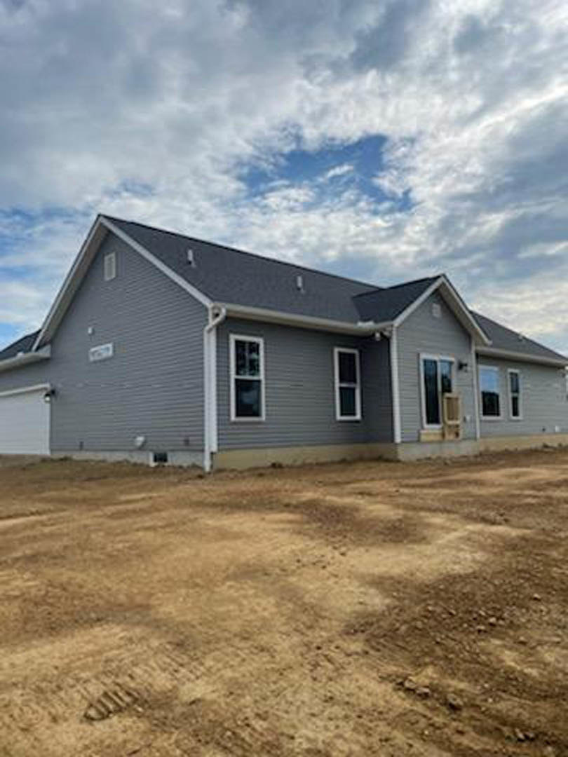 Partially built house with white framed windows, light siding, and exposed dirt field in foreground under blue sky with scattered clouds