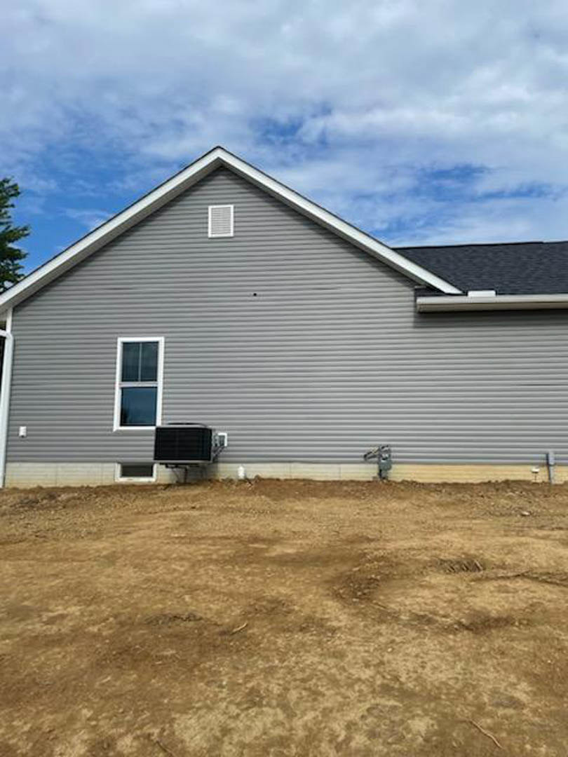 Grey-sided house with white-framed window, white vent, and exterior air conditioner unit, set beside a dirt field under a cloudy sky.