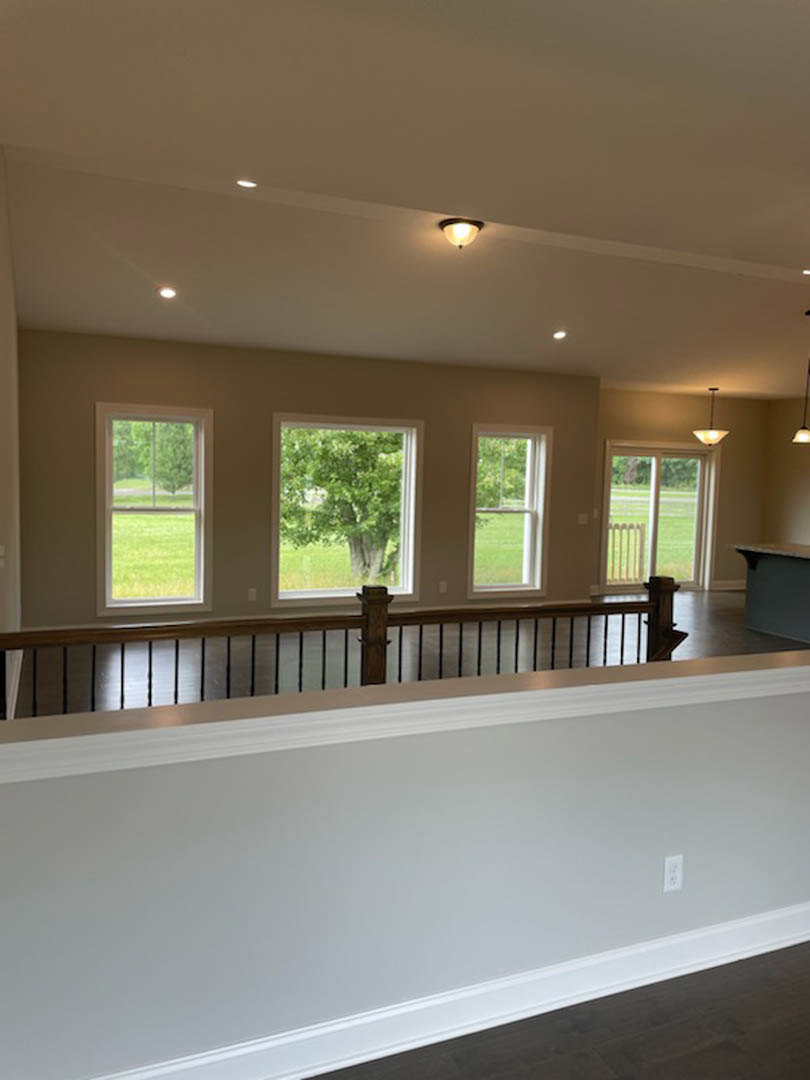 Open-concept room featuring a wooden staircase with black railing, large windows overlooking a green lawn and trees, white walls, and light hardwood flooring.