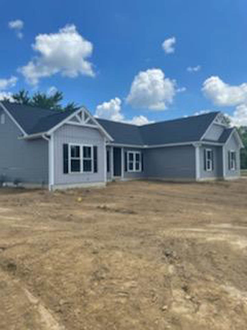 Partially built house with exposed framing and siding, set on a dirt lot under a blue sky with scattered clouds