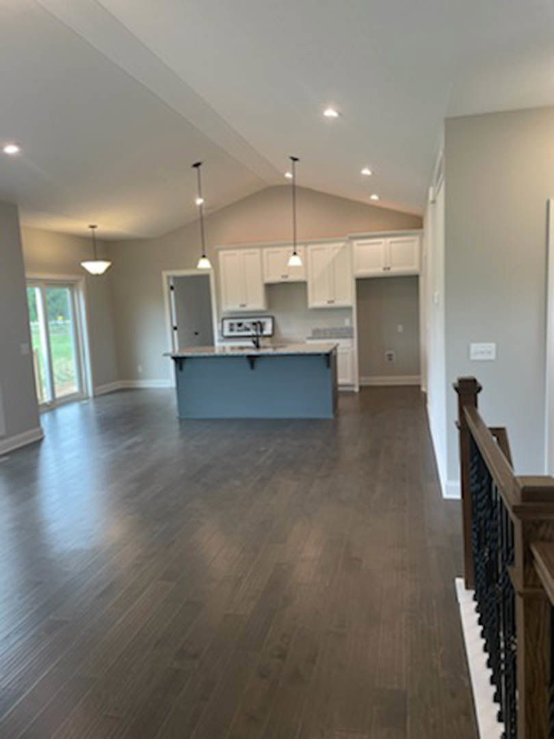 Open kitchen and living room with hardwood flooring, white cabinetry, wood shelving, and large window overlooking green lawn