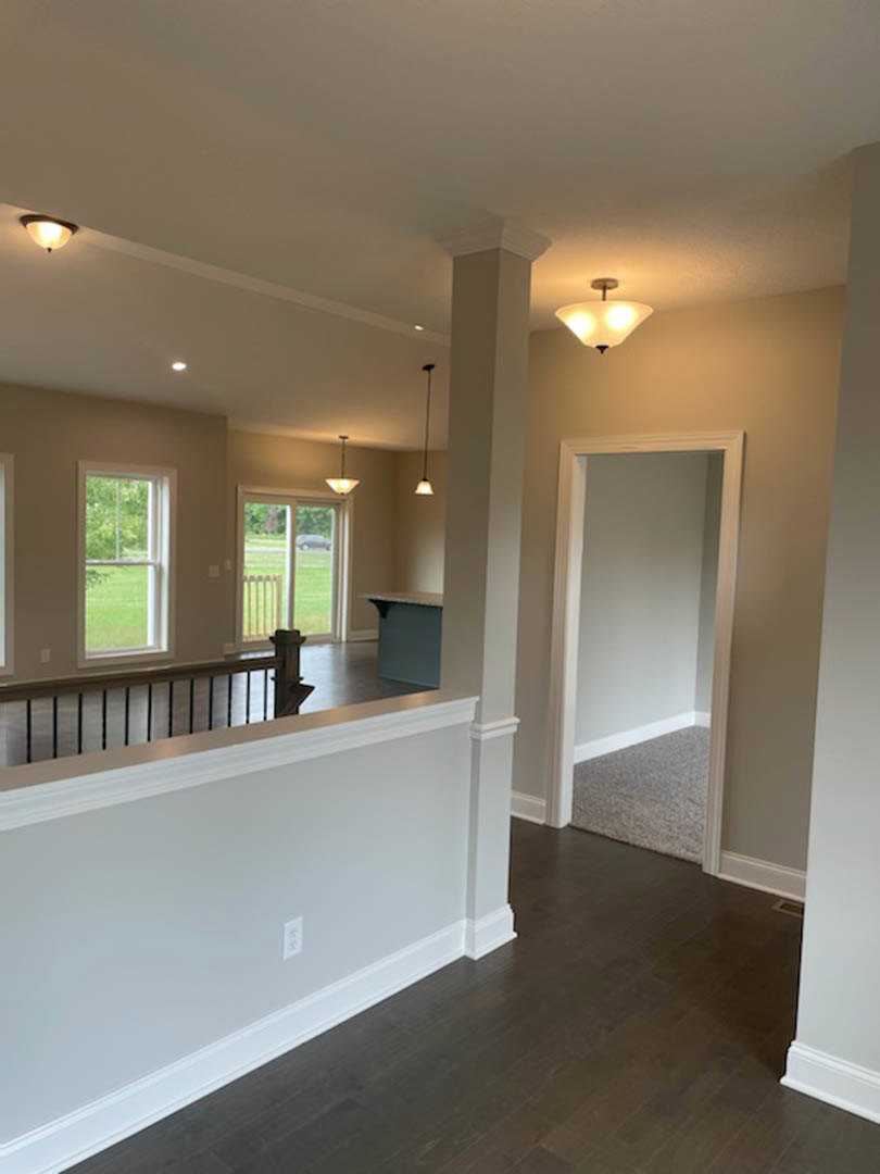 Hallway with white walls, wood flooring, staircase with white railing, door at the end, windows showing green lawn and parked car outside, ceiling light fixture illuminated