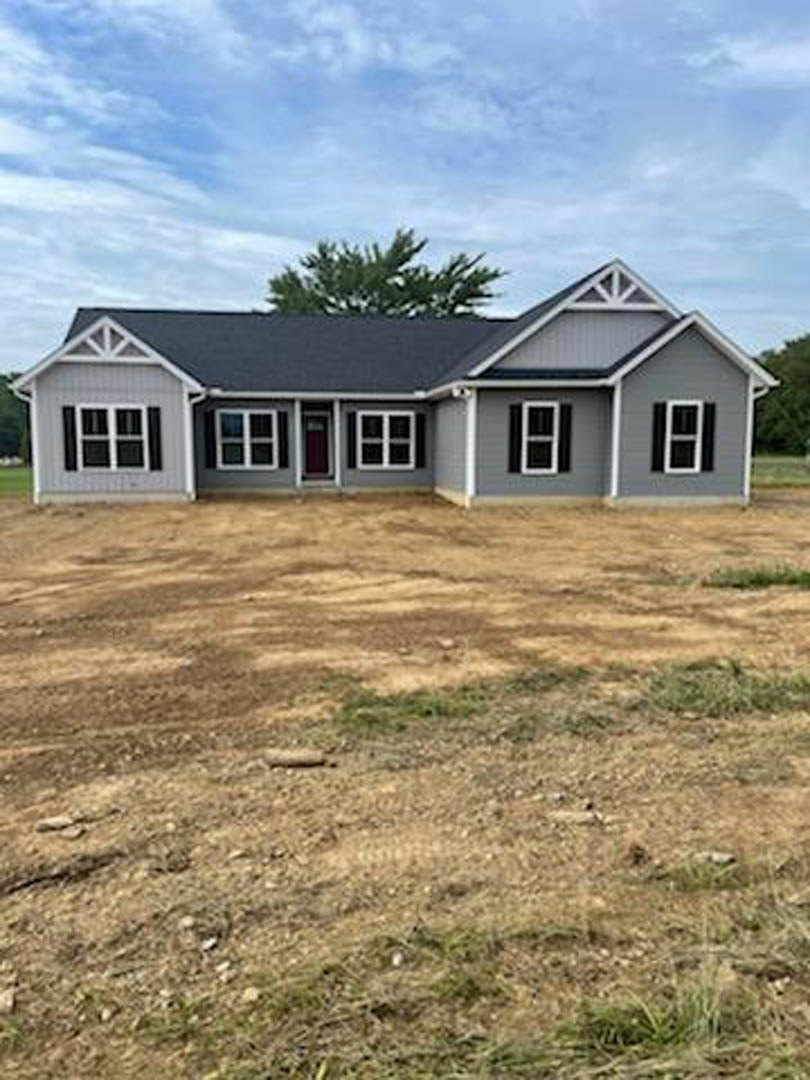 Two-story house with white-framed windows, gray roof, and a dirt field in the foreground; tree and cloudy sky in the background
