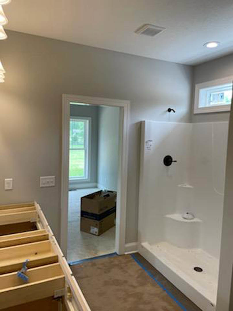 Bathroom with light-colored plaster walls, built-in bathtub, wooden shelf featuring a blue handle, white electrical outlet, walk-in shower with chrome shower head, cardboard box