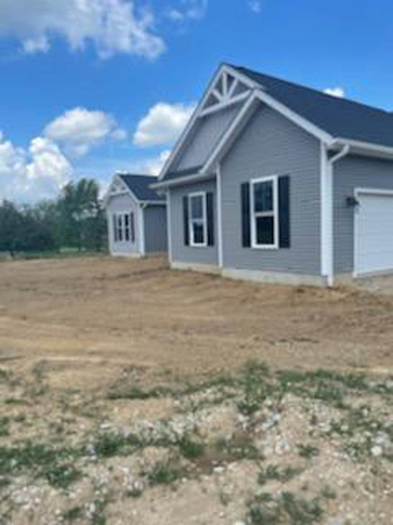 Partially built house with exposed framing, white siding, and large windows, surrounded by dirt lot under clear blue sky with scattered clouds