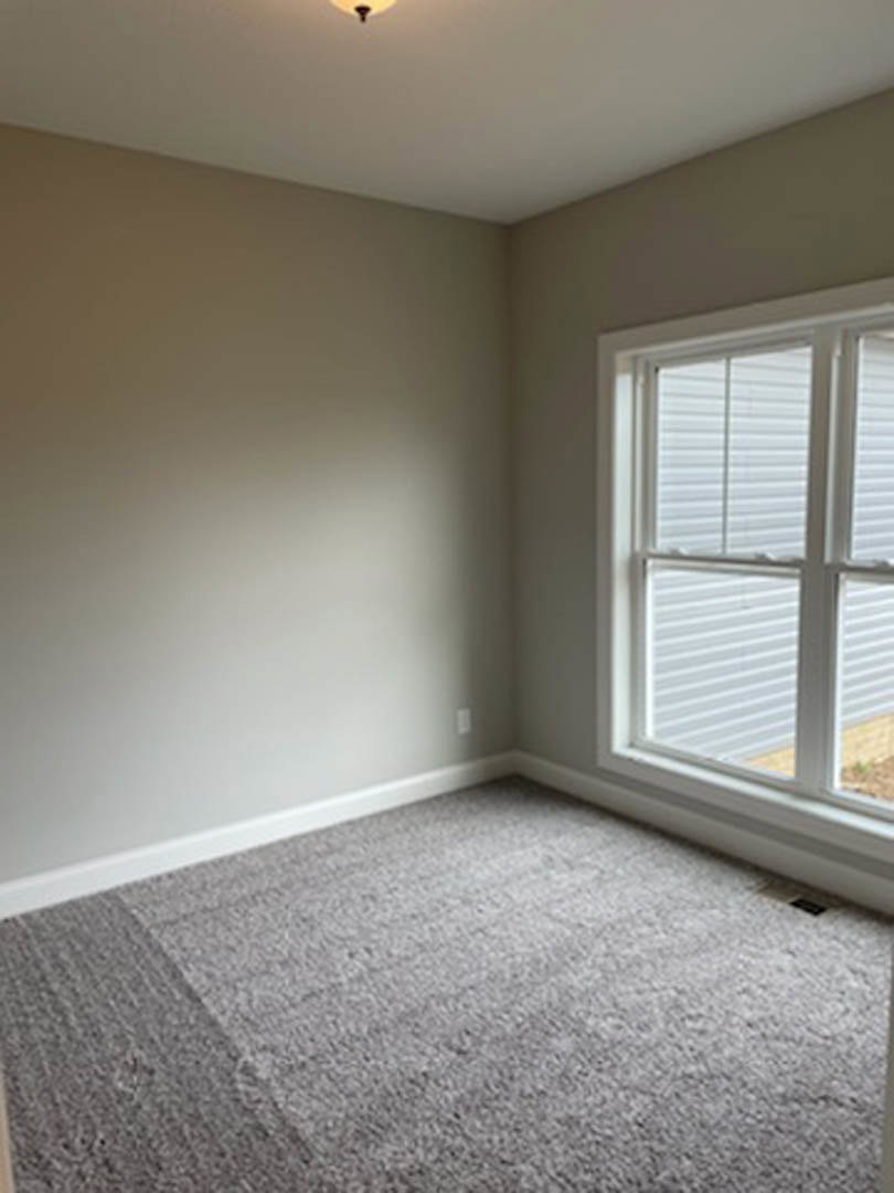 Sunlit room with white-framed window, grey carpet flooring, and neutral plaster walls