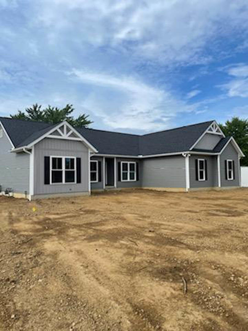 Modern single-story home with white exterior walls, large square windows, dirt landscaping in the foreground, palm tree and other trees in the background, under a partly cloudy