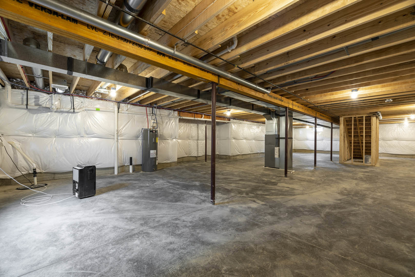 Wooden ceiling with exposed metal beam, concrete floor, white wall, visible ceiling pipe, and unfinished roof framing in a basement room