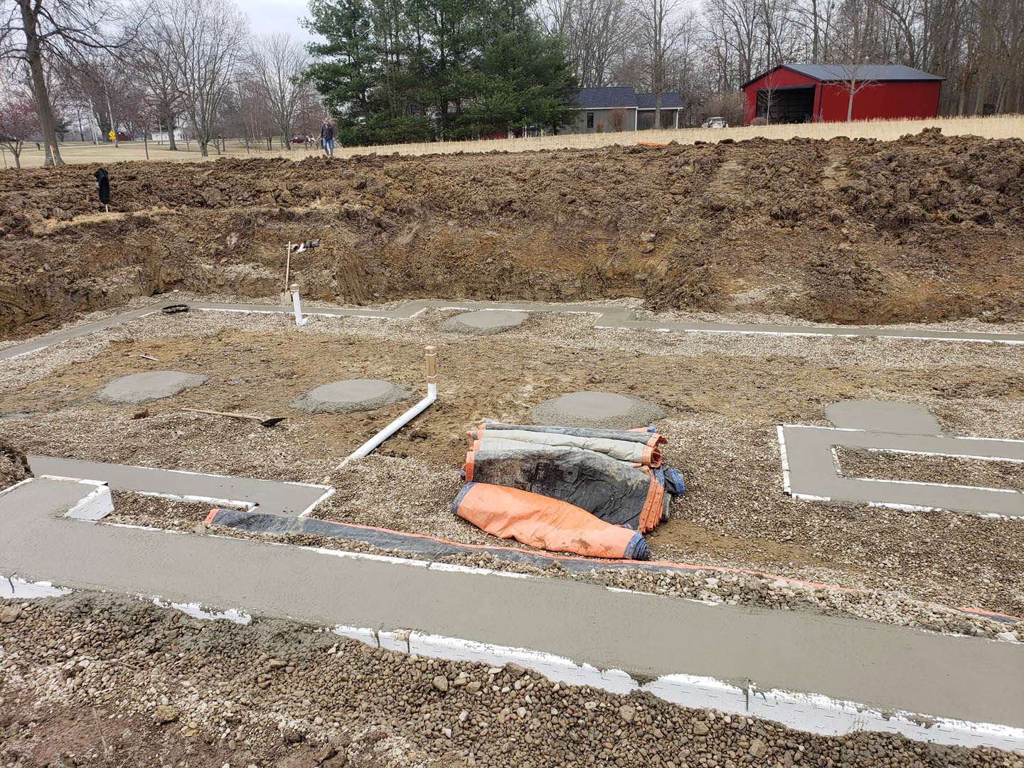Red barn with black door surrounded by trees, construction site featuring exposed soil, large hole, scattered dirty bags, orange bag, and puddle in the dirt foreground