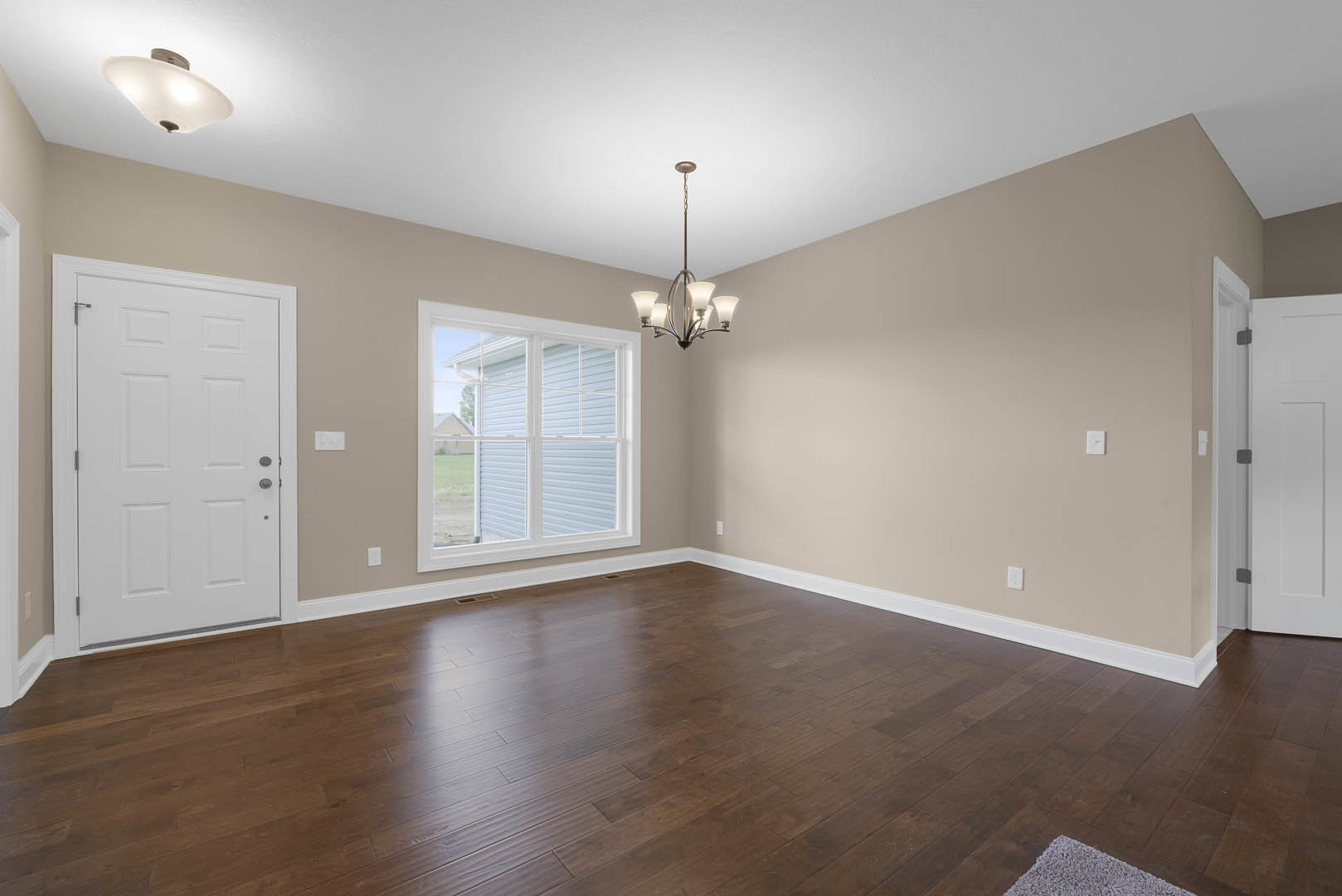 Hardwood floor room featuring a white door with silver handle, window overlooking neighboring house, and chandelier with white shade