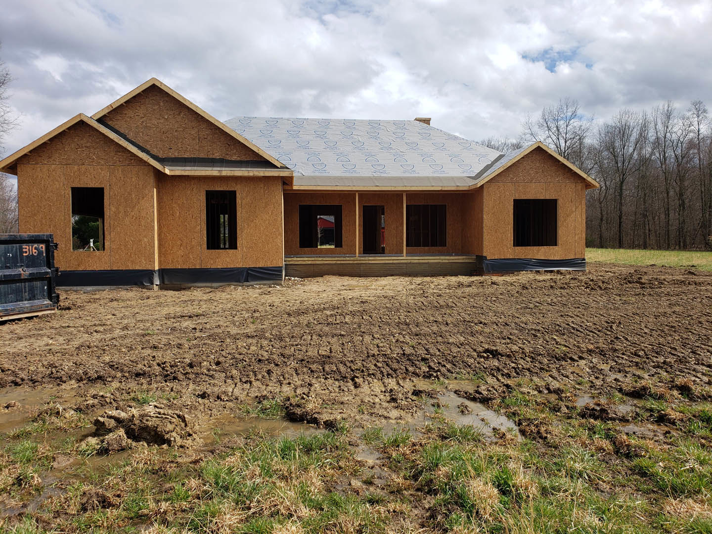 Partially built house with exposed framing and white tarp covering roof, surrounded by muddy field and patches of grass, black container near entrance, unfinished door visible