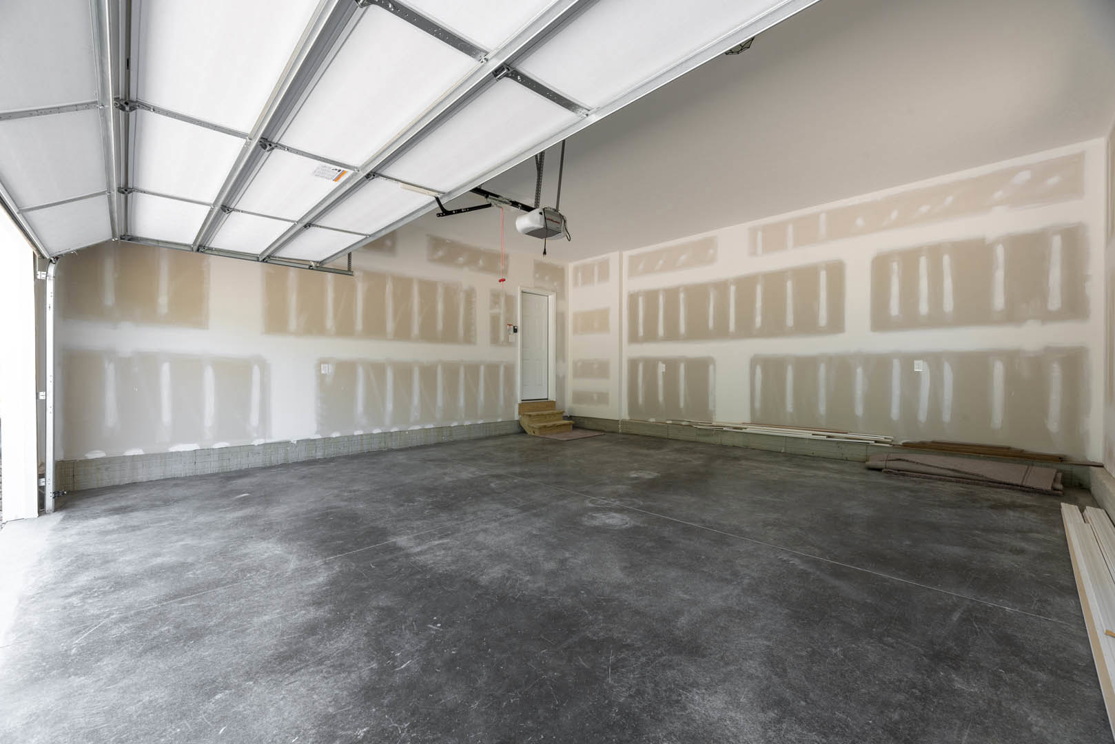 Concrete floor and white walls in an empty room, featuring a white door with black trim and handle, staircase visible in the background, daylight streaming in.