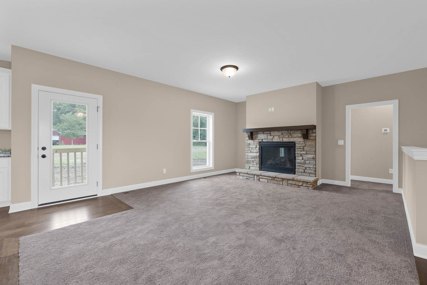 Living room with beige carpet, white-framed window, glass-front fireplace, ceiling light fixture, and door opening to view of red barn and tree.