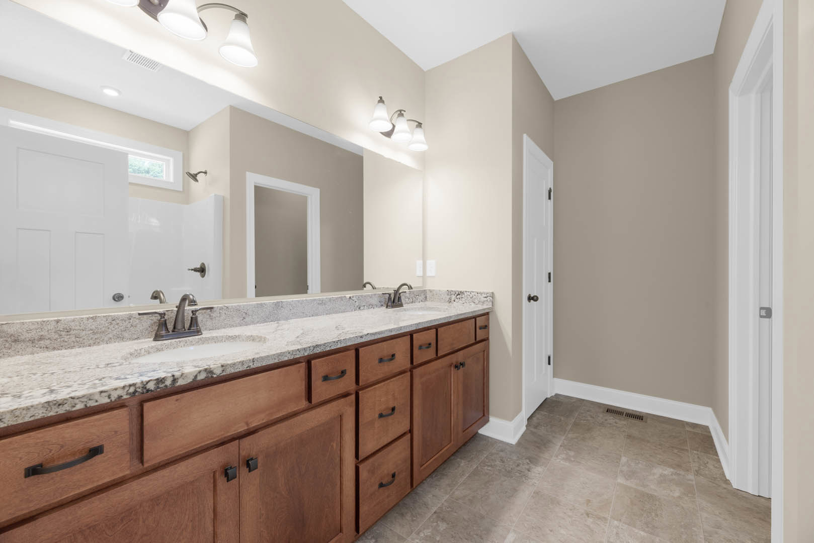Double vanity bathroom with white door frame, three-light fixture above mirrors, tile flooring, stone countertop, chrome faucets, and shaker-style cabinetry with drawers and