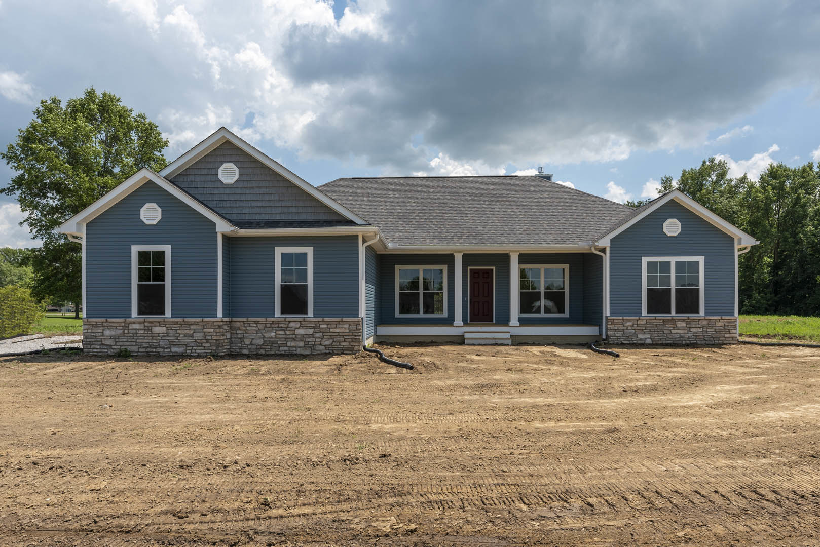 Blue exterior house with white-framed windows, red front door, and dark brown side door, set beside a dirt area with exposed pipe under a cloudy sky.