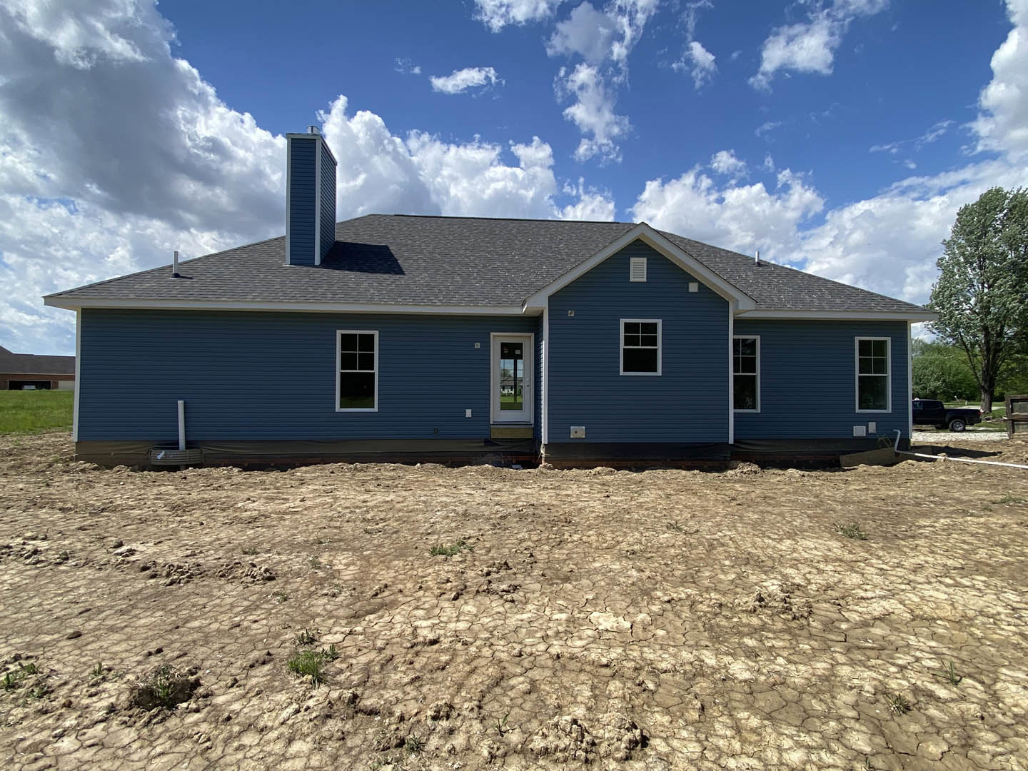 Blue house with white door and window, surrounded by cracked dirt field, leafy tree nearby, cloudy sky overhead