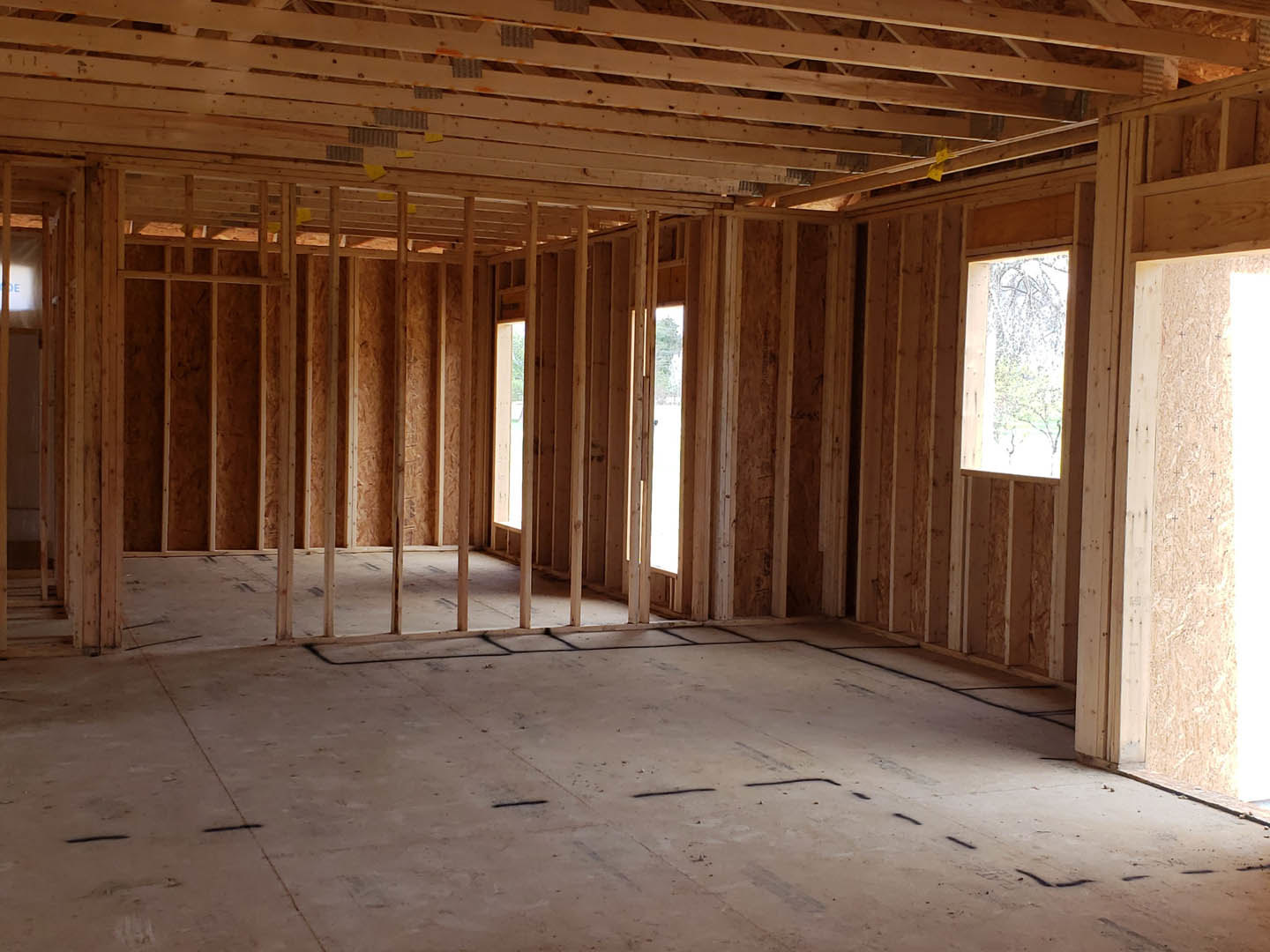 Living room with exposed wood ceiling beams, large windows framed in wood, light-colored plank flooring with black accent lines, and a wooden wall adjacent to a modern door.